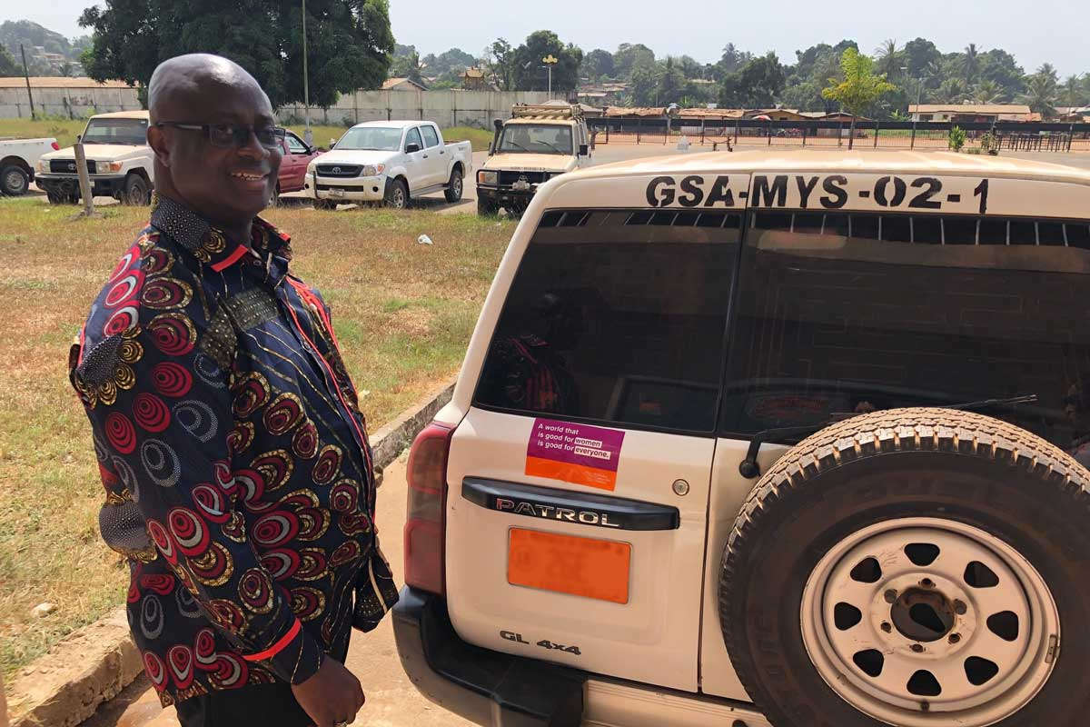 Liberia's Minister of Youth & Sports, Hon. Zeogar Wilson, affixes a WCW bumper sticker to his official vehicle
