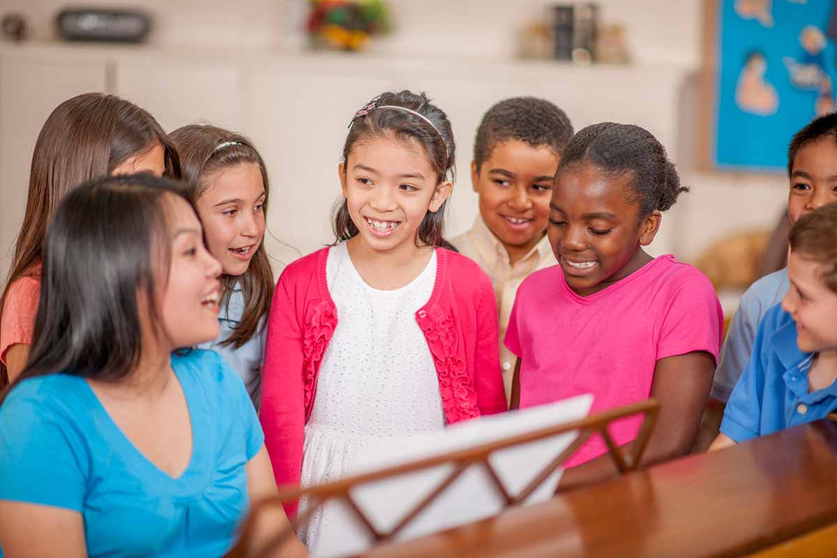 A diverse, happy group of kids stand around a piano singing