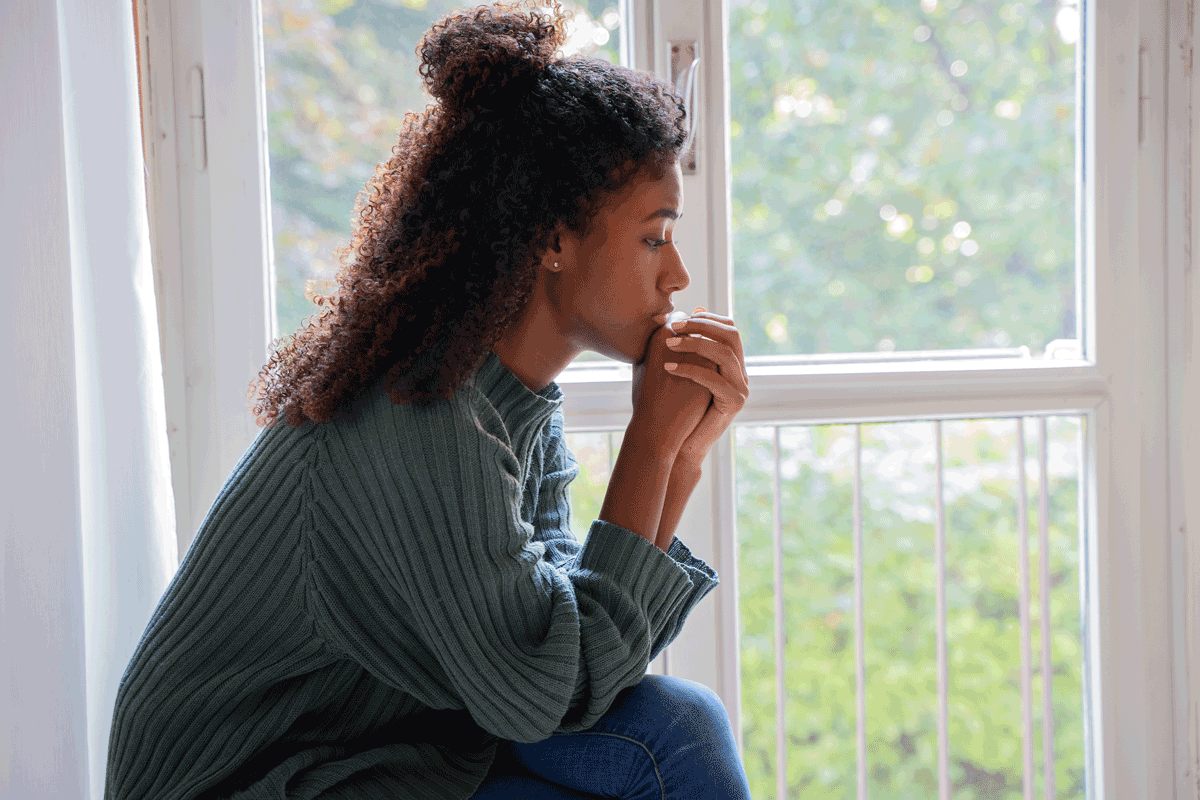Solemn young Black woman sits in front of window with light shining in