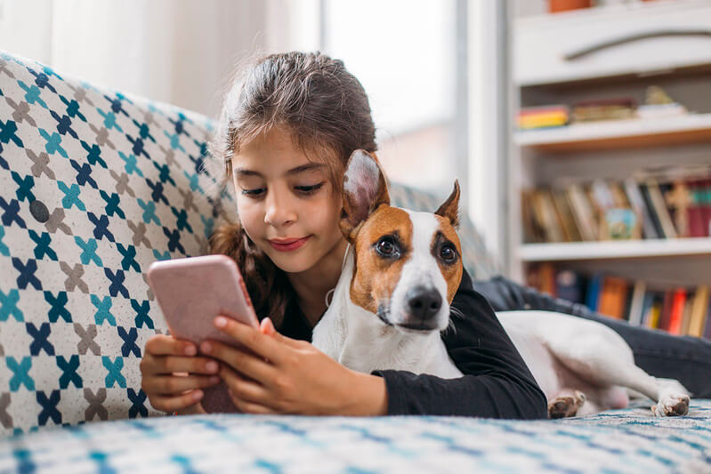 A young girl lying down on a sofa with her dog while using her phone