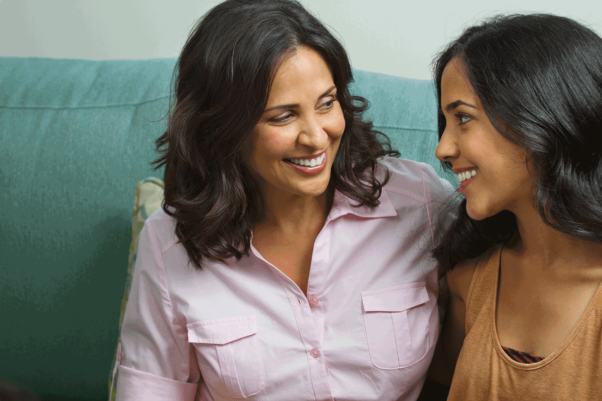 Latina woman and teen girl sit on couch and look at one another, smiling. 