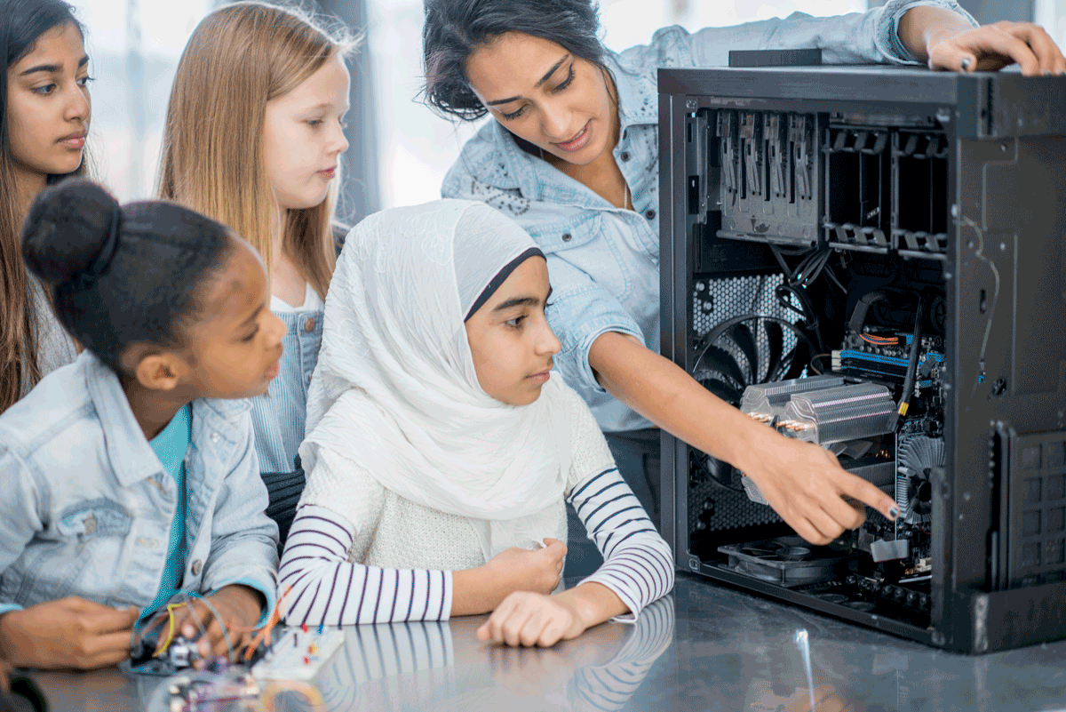 An adult woman teaches a diverse group of girls about computers in an informal learning setting.
