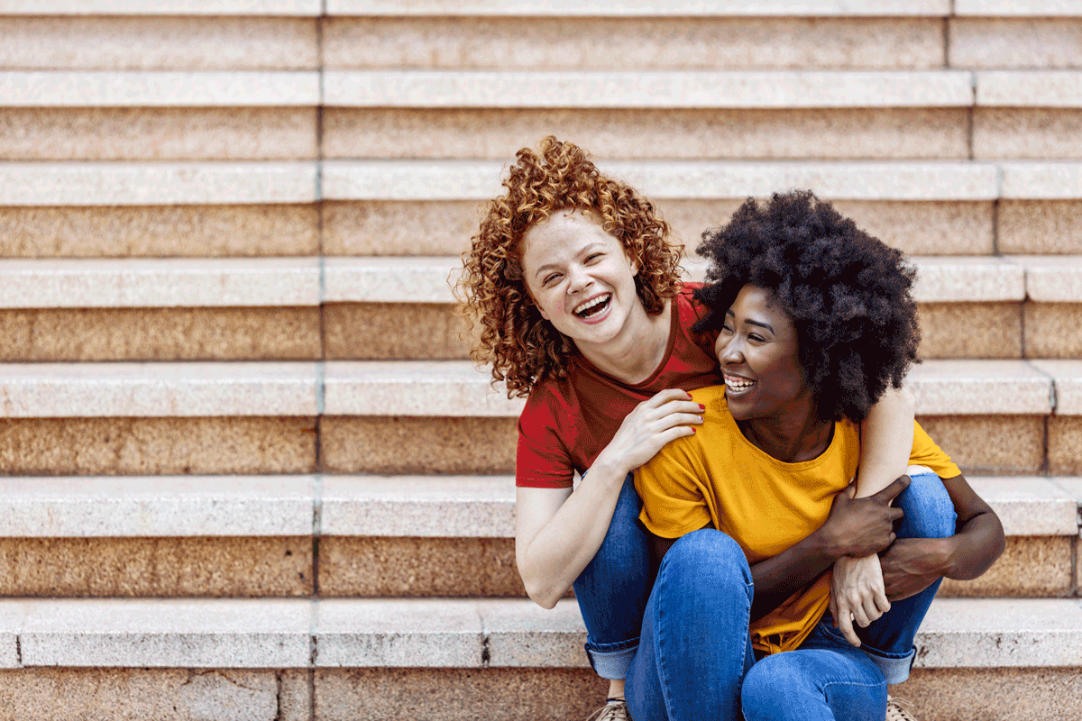 A biracial couple of teen girls smile while hugging outside.